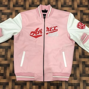 May include: A pink bomber jacket with white sleeves. The jacket features the text "Avi Rex USA" in red and white. The sleeves have a perforated texture. The jacket has a black zipper and white stripes on the cuffs and hem.