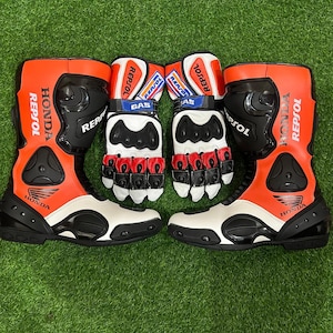 Puede incluir: Un par de botas de moto naranjas, blancas y negras con guantes a juego que presentan la marca Repsol y Honda. Las botas tienen paneles protectores negros y los guantes incluyen protectores de nudillos con logotipos Gas y Repsol.