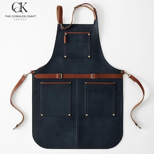 Puede incluir: Un delantal de cuero azul marino con correas y bolsillos de cuero marrón. El delantal tiene dos bolsillos delanteros grandes, un bolsillo en el pecho y correas ajustables. El texto "THE COBBLER CRAFT CRAFTED" está impreso en la parte superior izquierda.