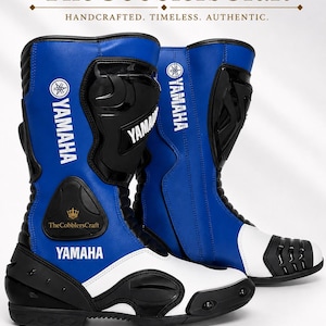 Puede incluir: Un par de botas de carreras de motos en azul, negro y blanco con el logotipo de Yamaha en texto blanco y una espinillera protectora negra. Las botas incluyen un refuerzo de talón negro, una puntera blanca y un parche circular TheCobblersCraft.