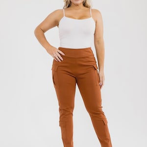 May include: A woman wearing a white spaghetti strap tank top and rust-colored cargo pants. The pants have multiple pockets and a high waist. The woman is wearing clear heeled sandals.