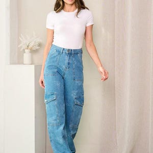 May include: A woman wearing a white short-sleeve t-shirt and light blue denim cargo pants. The pants feature a butterfly pattern and two cargo pockets on each leg. The woman is also wearing white sneakers.