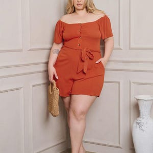 May include: A rust-colored off-the-shoulder romper with short sleeves, a button-up front, and a tie-waist. The model is holding a small woven handbag and wearing nude heels. A white vase is in the background.