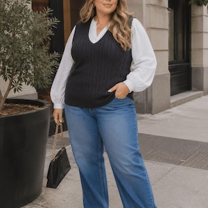 May include: A black cable-knit sweater vest layered over a white long-sleeve blouse with full sleeves, paired with blue denim jeans and a black handbag with a gold chain strap. The outfit is completed with black pointed-toe shoes.