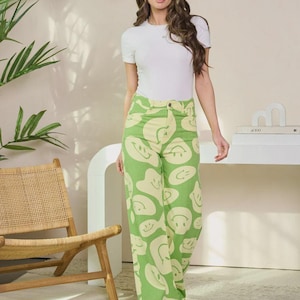 May include: A woman wearing a white short-sleeve top and wide-leg pants. The pants are light green with a pattern of off-white smiley faces. The background includes a wooden chair, a white table, and a plant.