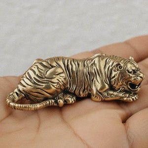 Könnte beinhalten: Kleine, detaillierte Tigerfigur aus Messing in Ruheposition. Der Tiger hat komplizierte Streifen und Gesichtszüge, mit leicht geöffnetem Maul. Die Figur ist etwa 7.6 cm lang.