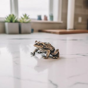 May include: A detailed bronze frog figurine with textured skin and realistic features. The frog is positioned on a white marble surface, showcasing its intricate design. The figurine is a decorative item, possibly for home decor or a collector's piece.