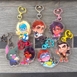 May include: A collection of seven acrylic keychains featuring various cartoon characters with colorful heart-shaped and bat-shaped clasps. The characters include a blonde hero, a person with a dog, a dark-haired figure, a bat creature, a red demon, a person with blue glasses, and a fire-wielding character.