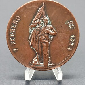 May include: A circular copper-toned commemorative medal featuring a relief of a figure holding a flag. The text 7 FEBRERO DE 1824 is embossed along the sides, and the name ORZALI B.Y C. is visible near the bottom edge of the central design.