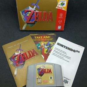 May include: A Nintendo 64 video game set for The Legend of Zelda: Ocarina of Time, including a gold cardboard box, a grey game cartridge, a Spanish instruction manual, a promotional flyer, and a white consumer information and precautions booklet.