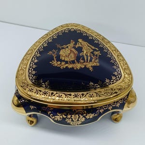 May include: A navy blue, heart-shaped trinket box with gold trim and feet. The lid features a gold illustration of two figures in a garden setting. The box has a decorative gold border and is likely made of porcelain.