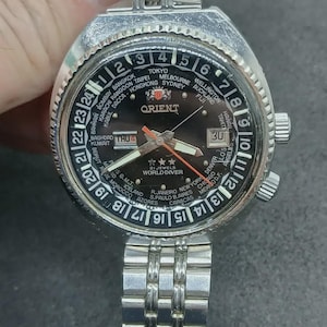 May include: A silver-tone Orient World Diver wristwatch with a black dial, featuring a 24-hour rotating bezel with global city names, a day-date window, luminous hands, an orange second hand, and a stainless steel link bracelet with a fold-over clasp.