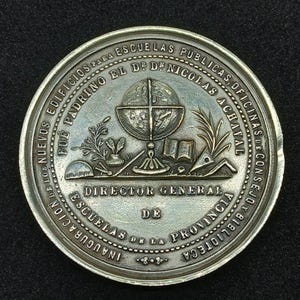 May include: A circular bronze-toned commemorative medal featuring a central relief of a globe, an open book, and botanical elements. The text reads: FUE PADRINO EL DR. NICOLAS ACHAVAL, DIRECTOR GENERAL DE ESCUELAS DE LA PROVINCIA, INAUGURACION DE LOS NUEVOS EDIFICIOS PARA ESCUELAS PUBLICAS, OFICINAS DE CONSEJO Y BIBLIOTECA.