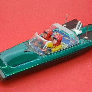 Könnte beinhalten: Ein grünes Spielzeugboot mit glänzender Oberfläche, ausgestattet mit einer transparenten Windschutzscheibe und zwei Figuren in roten Helmen und gelben Anzügen. Das Boot hat silberne Antennen und ein detailliertes Armaturenbrett. Das Spielzeug steht vor rotem Hintergrund.