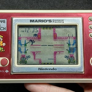 Könnte beinhalten: Ein tragbares elektronisches Nintendo Game & Watch Spiel mit dem Titel Mario's Cement Factory. Das Gerät verfügt über ein rot-weißes rechteckiges Gehäuse mit einem digitalen Bildschirm, der das Spielgeschehen anzeigt, Steuertasten und das Game & Watch Logo.