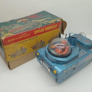 Könnte beinhalten: Vintage blaues Blechspielzeug-Raumschiff mit klarer Kuppel und roter Innenausstattung. Auf der Box steht "SPACE VEHICLE" und "Battery Operated with Floating Satellite". Das Spielzeug hat ein Retro-Design mit Illustrationen.