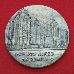 May include: A circular silver-toned metal medallion featuring a detailed relief of a grand building with ornate architectural columns and a flag on top. The text BVENUS AIRES ARGENTINA is embossed at the bottom, with the signature Roberti below it.