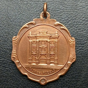 May include: A copper-colored medal with a detailed relief of a building, inscribed with "CASA DEL PUEBLO." The medal has an ornate, decorative border and a loop for hanging. The building features columns and multiple windows.
