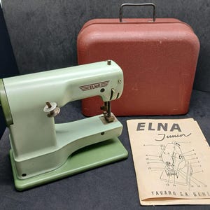 May include: A vintage light green Elna Junior sewing machine with a black hand crank, accompanied by a red carrying case with a handle and a beige instructional manual featuring a diagram and the text ELNA Junior TAVARO S.A. GENEVE.