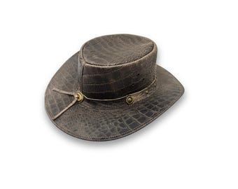 Può includere: Cappello in pelle marrone con motivo a trama simile alla pelle di coccodrillo. Il cappello presenta una corona arrotondata e un'ampia tesa. Una fascia decorativa con dettagli metallici circonda la corona. Un cordino in pelle con chiusura in metallo è attaccato lateralmente.