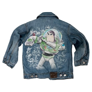 Puede incluir: Chaqueta vaquera azul con un gran gráfico de Buzz Lightyear de Toy Story en la espalda. La chaqueta tiene una parte delantera abotonada, dos bolsillos en el pecho y un cuello clásico. El gráfico de Buzz Lightyear es verde, blanco y morado.