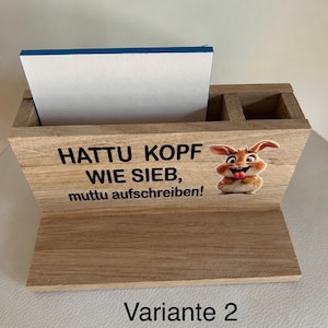 Könnte beinhalten: Ein hellbrauner Schreibtisch-Organizer aus Holz mit zwei Fächern und einer flachen Basis. Die Vorderseite zeigt eine Cartoon-Hasengrafik und den schwarzen Text: HATTU KOPF WIE SIEB, muttu aufschreiben! Ein weißer Notizblock steckt im Fach.
