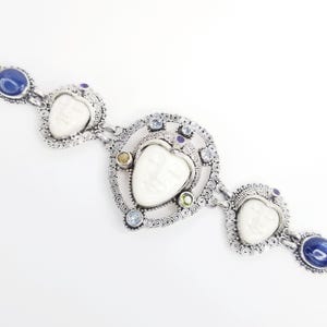 May include: A silver bracelet featuring three carved white face cameos set in intricate filigree frames. The design is accented with round blue, yellow, and green gemstones, including deep blue cabochons at the ends of the link chain structure.