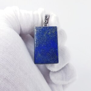 May include: A rectangular pendant featuring a deep blue lapis lazuli stone with natural gold-colored pyrite inclusions. The stone is set in a simple metal frame with a small, circular silver-toned bail at the top for attaching to a chain or cord.