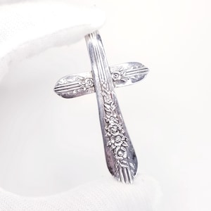 May include: A silver-toned metal cross pendant crafted from a repurposed spoon handle. The surface features an embossed floral pattern with detailed roses and leaves along the vertical stem and horizontal arms, showcasing a polished metallic finish.