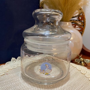 Könnte beinhalten: Ein klares Glas mit Deckel. Das Glas hat ein rosa-weißes Design am Rand und eine Cartoon-Gans mit Jungen auf der Vorderseite. Das Glas steht auf einer beigen Oberfläche.