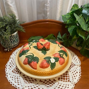 Könnte beinhalten: Eine cremefarbene Keramik-Kuchenform mit Deckel, verziert mit roten Erdbeeren und grünen Blättern. Die Form hat einen gewellten Rand. Der Deckel hat einen Griff aus zwei Erdbeeren. Sie steht auf einer weißen Häkelspitze.