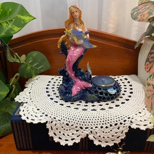 May include: A mermaid figurine with a pink tail and long blonde hair, holding a blue dolphin. The figurine sits atop a blue base with a tealight holder. The mermaid is on a white crocheted doily, placed on a blue book.