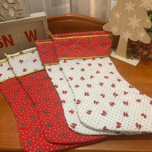 Peut inclure: Chaussettes de Noël à motifs rouges et blancs. Les chaussettes ont un dessus rouge à pois blancs et une bordure dorée. Le corps de la chaussette est blanc avec des motifs en forme de nœuds rouges. Un panneau en bois avec "SNOW" en arrière-plan.