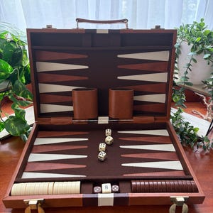 May include: Backgammon set, open and ready for play. The playing surface is brown, white, and tan. The wooden case has a handle and metal clasps. Includes dice, game pieces, and leather dice cups. The game is open and ready to play.
