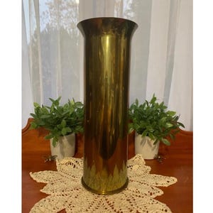 Puede incluir: Un jarrón alto y cilíndrico de latón con la parte superior ensanchada. El jarrón mide aproximadamente 30 cm de alto y se apoya sobre un tapete de ganchillo color crema. Dos pequeñas plantas en maceta flanquean el jarrón, añadiendo un toque de vegetación.