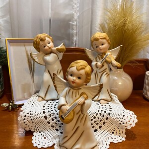 Puede incluir: Tres figuras de ángeles de cerámica con cabello rubio y túnicas blancas. Un ángel toca un laúd, otro un violín y el tercero una trompeta. Cada uno tiene alas doradas. Están dispuestos sobre un tapete de encaje blanco, con un cuadro enmarcado y un jarrón en el fondo.