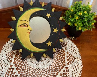 May include: A hand-painted wooden mirror in the shape of a crescent moon and sun. The moon is yellow with a smiling face, and the sun is black with yellow and gold accents. The mirror is displayed on a white lace doily.
