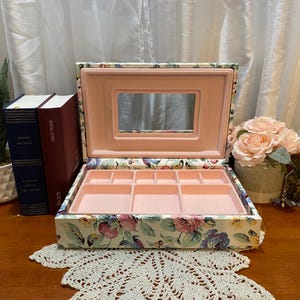 May include: Open jewelry box with a floral pattern, featuring a pink interior and mirror. The box is on a wooden surface, with books and a potted plant in the background. The box is designed to store jewelry.
