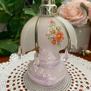 May include: A vintage, iridescent pink glass perfume bottle with a floral design. The bottle has a frosted glass shade with a flower pattern and hanging crystal accents. The top is silver-toned metal.