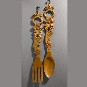 Può includere: Viene mostrata una coppia di utensili da cucina decorativi in legno, una forchetta e un cucchiaio. Gli utensili sono scolpiti con disegni floreali e di foglie e sono di colore marrone chiaro. Sono appesi a una parete grigia.