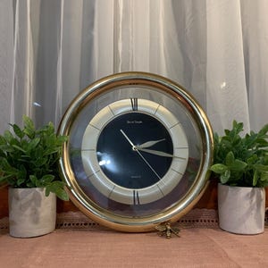 Peut inclure: Une horloge murale ronde dorée avec un cadran noir et des aiguilles dorées. L'horloge a un couvercle en verre transparent et est entourée d'une lunette dorée. L'horloge est flanquée de deux petites plantes en pot et d'une abeille décorative.
