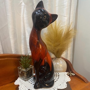 May include: A ceramic cat ornament with a glossy finish. The cat is predominantly black with red and brown detailing. It has a long, slender body and is posed in a seated position. The ornament is a decorative item.