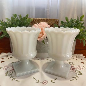 May include: A pair of white milk glass vases, each with a scalloped rim and a pedestal base. The vases are set on a floral embroidered tablecloth. Greenery and a pink flower are visible in the background. The vases have a vintage style.