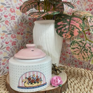 Può includere: Un vaso di ceramica bianco con coperchio rosa, decorato con un cigno e un motivo a cestino floreale. Un'ape decorativa e un fiore rosa sono su una fetta di legno. Un vaso bianco contiene una pianta con foglie rosa e verdi.