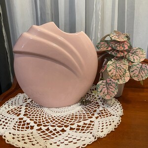 May include: A pale pink ceramic vase with a distinctive, curved design. The vase rests on a white, crocheted doily. A small potted plant with pink and green leaves is to the right of the vase.