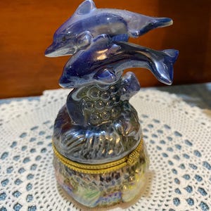 May include: A ceramic trinket box featuring two blue dolphins leaping above a textured base. The box has a gold-colored rim and a pearlescent finish, creating a shimmering effect. The dolphins are a darker shade of blue.