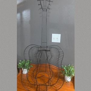May include: A black metal guitar-shaped decorative piece. The guitar is made of wire and sits on a wooden surface. Small potted plants are placed on either side of the guitar. The background is a gray wall.