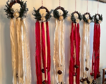 Puede incluir: Una colección de coronas decorativas colgantes. Cada corona está hecha de ramitas y vegetación, adornada con flores blancas y largas cintas fluidas en crema y rojo. Adornos marrones en forma de bellota cuelgan de las cintas, suspendidos de una rama de madera natural.