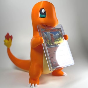 May include: An orange Charmander figurine holding a Pokémon trading card in a protective case. The figurine has white claws, a yellow belly, and a tail with a red and yellow flame. The card features the name "Charmander".