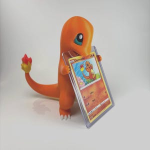 May include: An orange Charmander figure holding a Charmander trading card in a protective case. The figure has a flame-shaped tail with red and yellow accents. The card features a colorful illustration of Charmander.